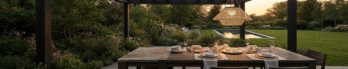Outdoor dining area under a wooden pergola, featuring a large natural rattan pendant light hanging above a set wooden table at dusk.
