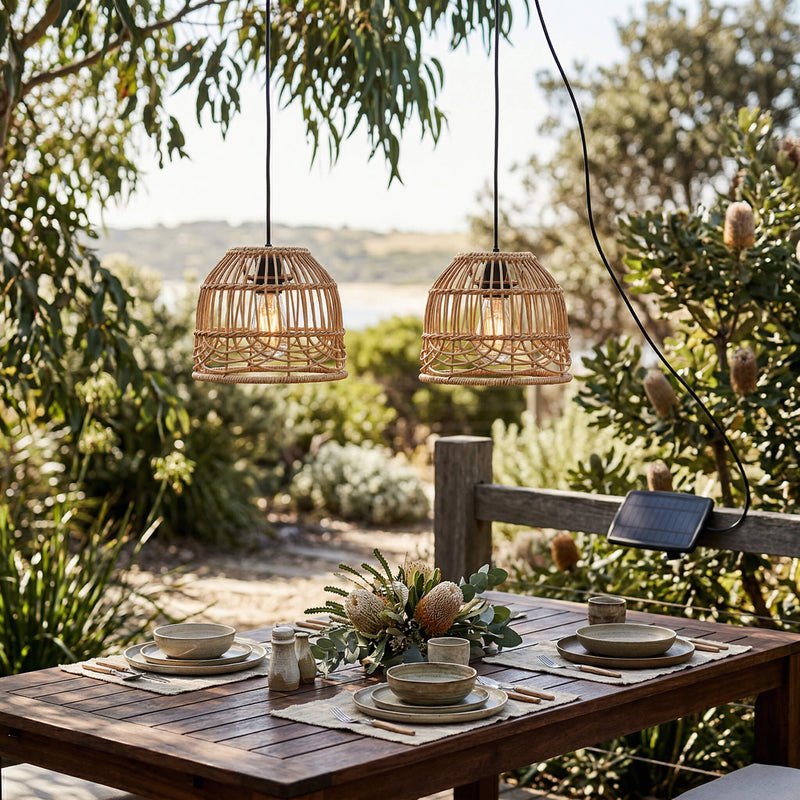 Outdoor dining setup with wicker pendant lights, tableware, and a solar panel in a garden setting.