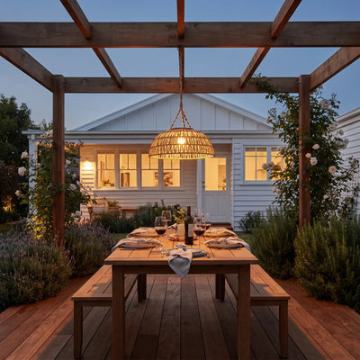 Haiku PT0901 solar rattan pendant hanging over wooden pergola dining table at dusk, casting warm glow on coastal garden with ocean view.