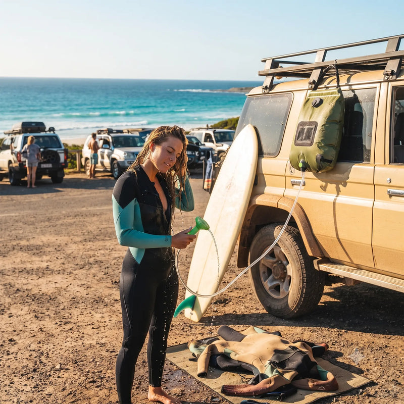 Surfer rinses wetsuit with solar shower bag hung on 4WD at beach; ocean and parked vehicles in background.