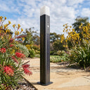 Black outdoor bollard light in a garden setting with plants and a pathway.