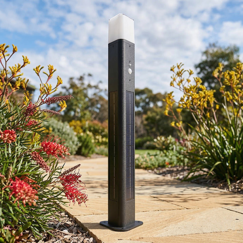 Black outdoor bollard light in a garden setting with plants and a pathway.