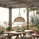 A handwoven PT0901 Kiku Solar Rattan Hanging Lantern glowing warmly over an outdoor wooden dining table set for dinner under a pergola at dusk.