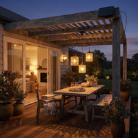 Outdoor patio dining setup under a wooden pergola at dusk, with a table, chairs, hanging woven pendant lights, a bench, potted plants, and a house interior visible through sliding glass doors.