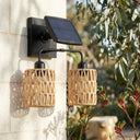 A modern solar-powered outdoor wall sconce with two woven rattan shades and a black solar panel, mounted on a light stone wall with green foliage in the background.