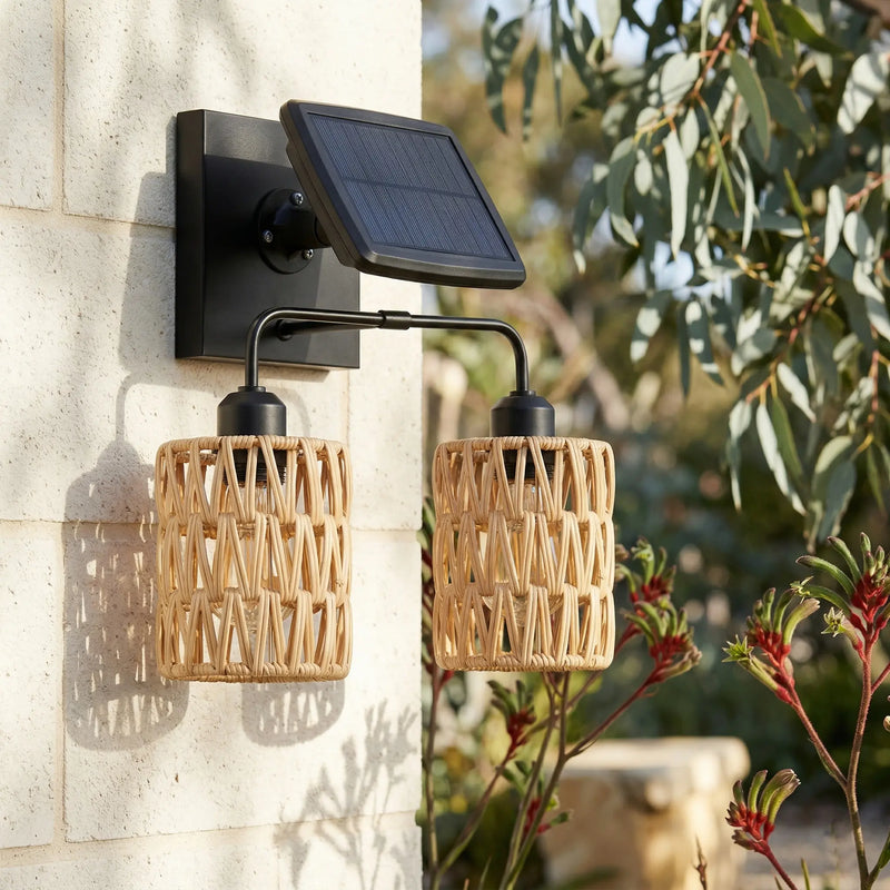 A modern solar-powered outdoor wall sconce with two woven rattan shades and a black solar panel, mounted on a light stone wall with green foliage in the background.