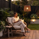 Woman reading on rattan chair beside LunaBeam PT0908 floor lamp — 148cm solar-powered woven shade casts warm glow on wooden deck, ideal for patio relaxation with zero wiring and IP54 weather resistance.