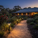 A landscaped garden pathway at dusk leading to a wooden house, illuminated by several black solar post lights with warm white glowing shades.