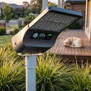 T008 smart solar street light with LED battery display showing 100% power and M3 mode, mounted on a pole in a residential garden with a dog resting on the deck in the background, highlighting real-time battery monitoring.