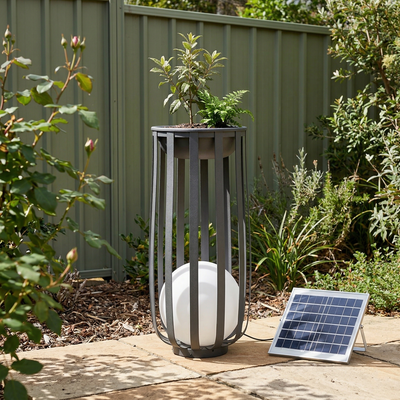 A solar-powered outdoor plant light and planter in a garden setting, featuring a tall dark gray metal cage with a glowing white globe lamp inside, alongside a small solar panel on a stone patio in front of a green fence.