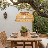 Dining table set outdoors with a wicker pendant light, white wall, and greenery.