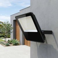 A sleek black E017EI Fano LED Floodlight mounted on a modern grey exterior wall, showcasing its minimalist design and bright illumination against a contemporary home facade with wooden accents.