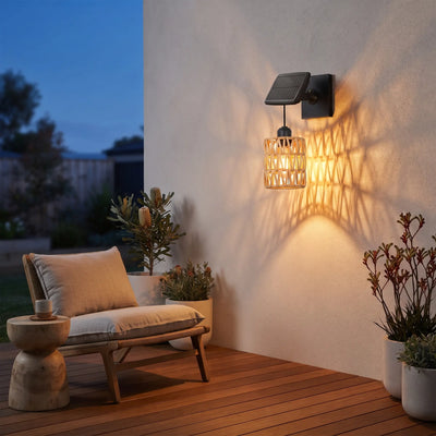 Haiku PT0909 solar rattan wall light mounted on white stucco wall at dusk, casting warm woven shadow patterns beside wooden deck chair and potted native plants — cozy alfresco ambiance with IP54 weatherproof design.
