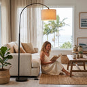 Woman reading a book in a cozy living room with a large window, floor lamp, and decorative items.