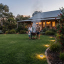 A family gathers around a smoking charcoal grill on a large green lawn in front of a house at dusk. Solar pathway lights illuminate the garden beds and lawn, and smoke rises from the grill.