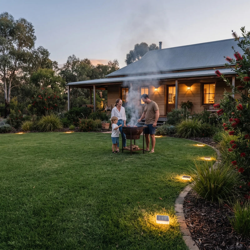 A family gathers around a smoking charcoal grill on a large green lawn in front of a house at dusk. Solar pathway lights illuminate the garden beds and lawn, and smoke rises from the grill.