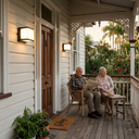 An elderly couple sits on a wooden porch reading a newspaper, with a small table between them. The porch has white siding, a wooden door, wall-mounted lights, and a view of lush tropical greenery beyond.
