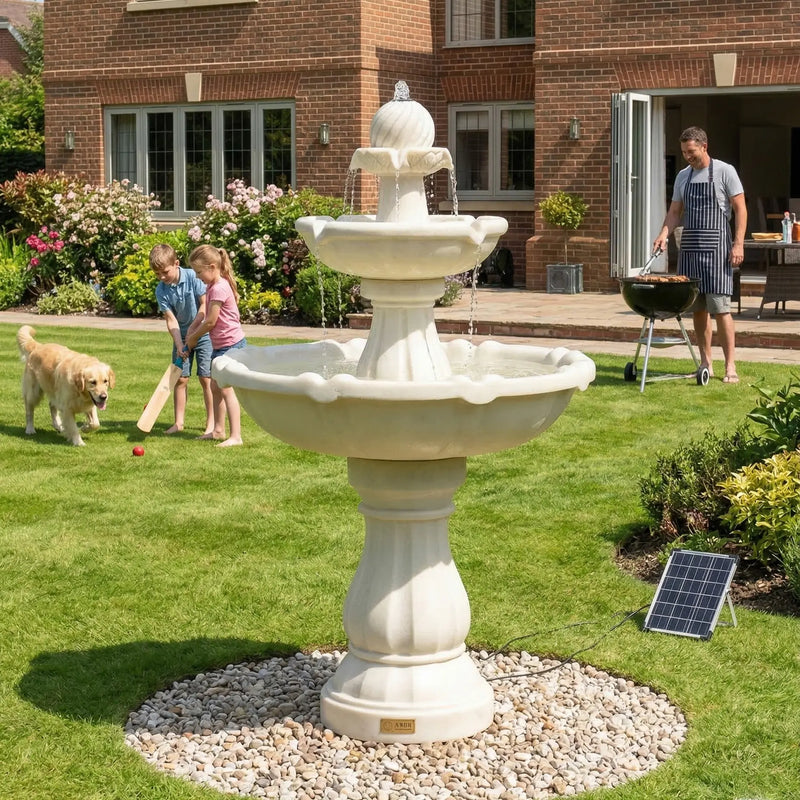 OUTWAY F001 two-tier solar garden fountain on a green lawn with children playing cricket and a golden retriever while a father barbecues in the background of a brick house