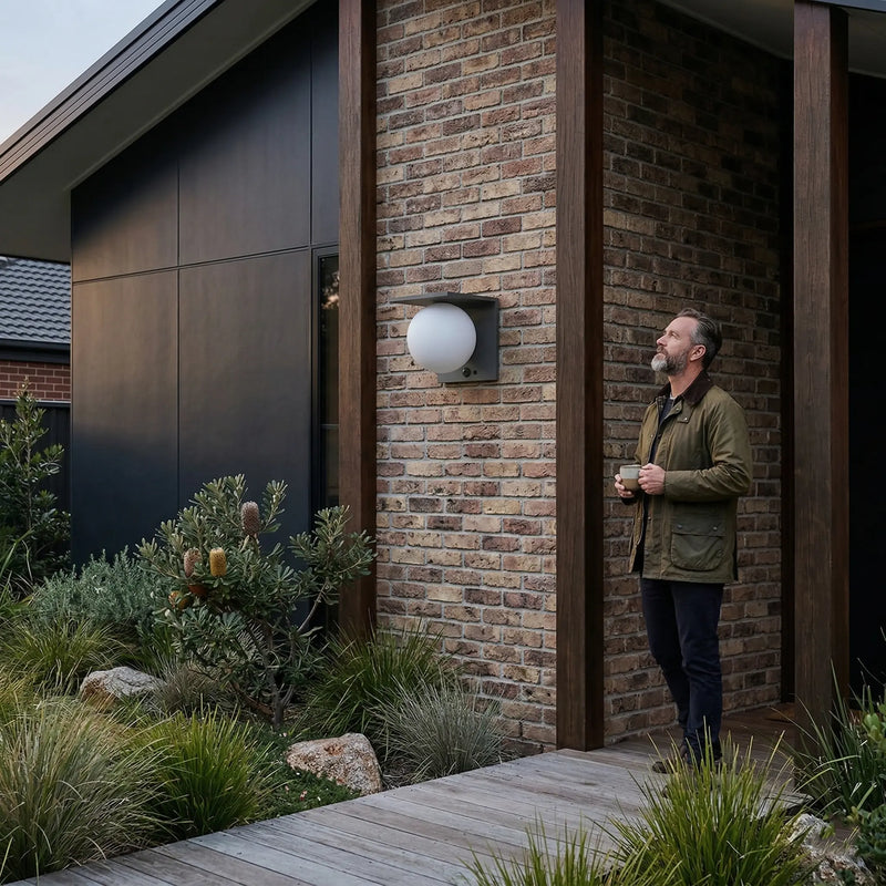 an holding coffee cup standing on timber deck outside modern brick home at dusk, looking up at Lumora Solar PT0720 globe wall light mounted on brick pillar, native Australian garden in foreground