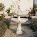 Two-tiered solar-powered angel fountain in ivory white resin, featuring a cherub statue atop cascading basins, set on gravel beside lavender and outdoor seating — ideal for elegant garden centers and tranquil patio ambiance.