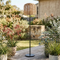 The 148cm PT0908 Solar Rattan Floor Lamp standing on an outdoor wooden deck surrounded by lush greenery and a stone wall, showcasing its natural aesthetic and tall, sculptural design.