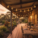 Solar string lights drape across a wooden veranda at sunset, casting warm glow over dining table and native garden beds.