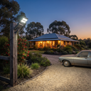 A rustic log cabin-style house with a blue metal roof, warmly lit windows, and solar panels on the roof, set at dusk with a gravel driveway, garden plants, a wooden post with a lit outdoor light, and part of a silver car in the foreground.