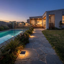 Modern outdoor stone walkway beside a swimming pool at dusk, with solar ground lights illuminating the path and landscaping in front of a contemporary house.