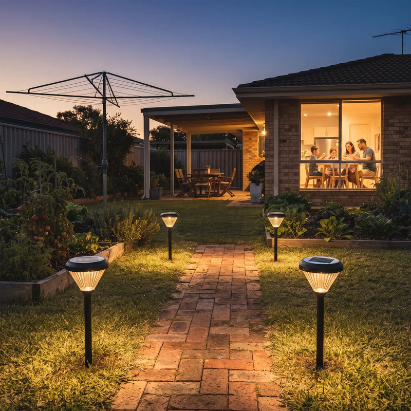 Family dines indoors as solar path lights guide through brick walkway flanked by raised veggie beds and clothesline.