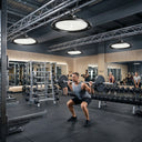 Outway high bay LED lights mounted on industrial ceiling trusses in a commercial gym, brightly lighting a man performing barbell squats with other gym members in the background.