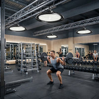 Outway high bay LED lights mounted on industrial ceiling trusses in a commercial gym, brightly lighting a man performing barbell squats with other gym members in the background.