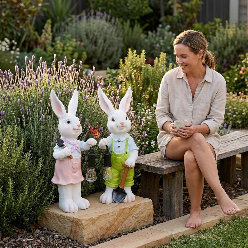 Woman sitting on a wooden garden bench smiling at Lumi A002 boy and girl rabbit solar ornament pair displayed among lavender and rosemary in an Australian garden with a monarch butterfly