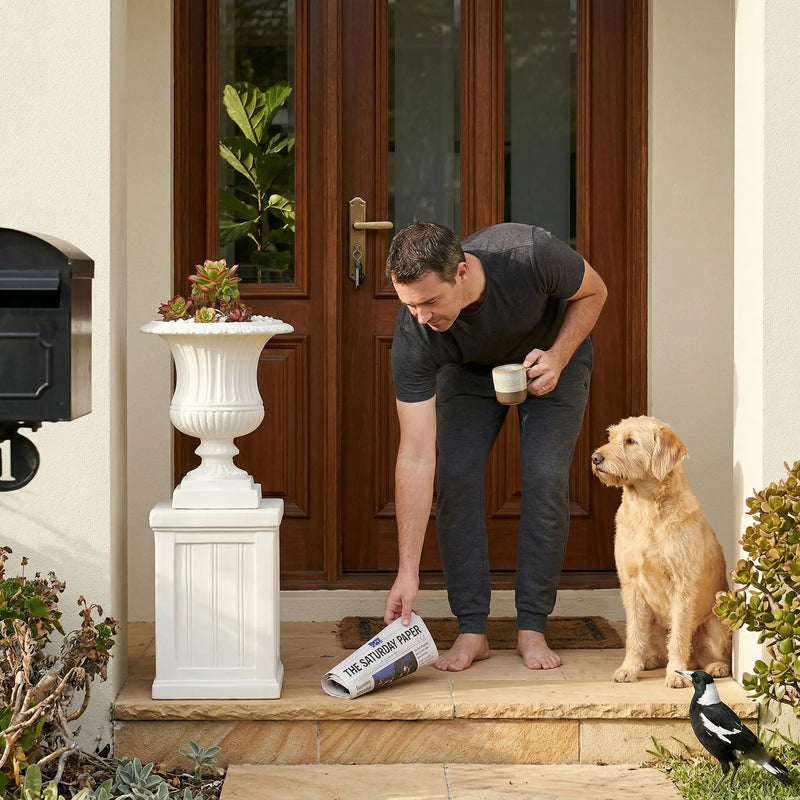 Man picking up morning newspaper at front door with white OUTWAY Flora Roman planter holding succulents on pedestal, golden retriever beside him — F004-F006-flora-combo-entrance-morning-lifestyle