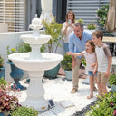 Family of four with father and two excited children pointing at OUTWAY F001 white two-tier solar garden fountain surrounded by colourful potted plants while mother photographs the moment