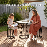 Two women sitting at a small round table on a wooden deck with plants and a white wall in the background.