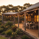 String lights illuminate a rustic timber deck beside a weatherboard home, with lush native plants and outdoor seating under twilight sky.