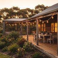 String lights illuminate a rustic timber deck beside a weatherboard home, with lush native plants and outdoor seating under twilight sky.