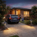 Rivix PT0723 solar bollard lights illuminating a residential driveway at dusk, with a woman unloading groceries from a Volvo SUV while children are visible reading inside a warmly lit brick home.