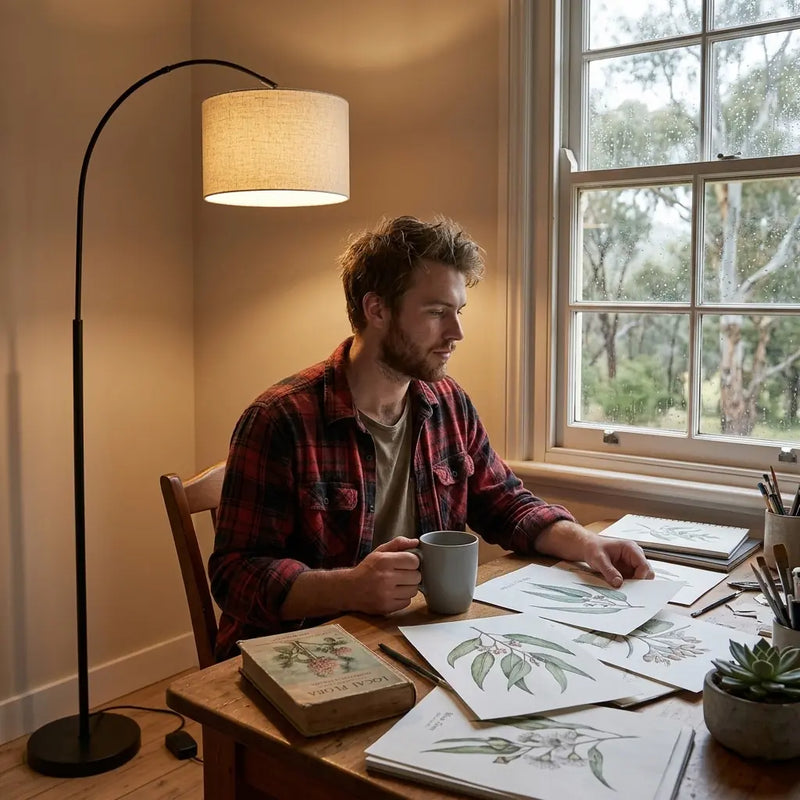 Man sitting at a table with art supplies and a cup, near a floor lamp and window with natural light.