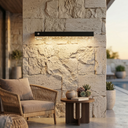 Outdoor setting with a stone wall, wicker chair, and table with plants, illuminated by a modern wall light fixture.