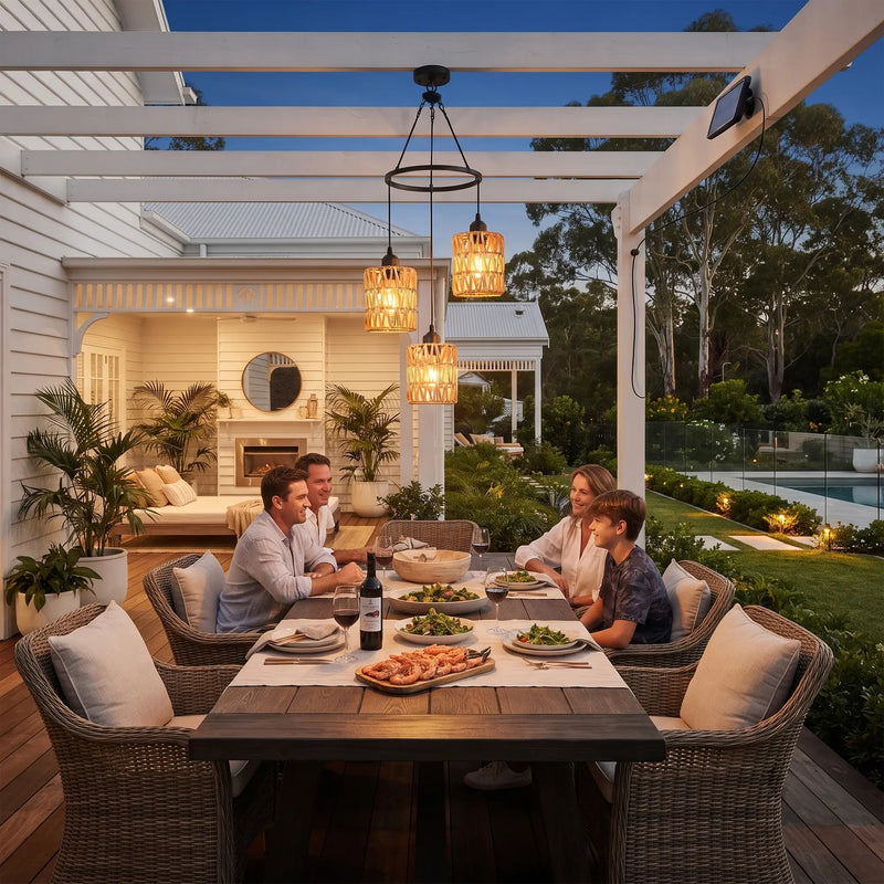 A family of four shares a meal at an outdoor wicker dining table under a white pergola at dusk. The scene is set on a backyard deck with a swimming pool, lush green landscaping, and a white house in the background.