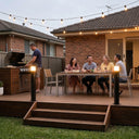 People sitting around a table on a backyard deck with string lights and a barbecue.