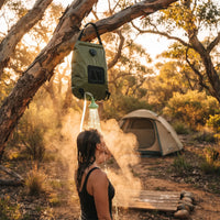 Woman showers under tree using solar-heated water bag; steam rises as tent glows softly in golden-hour forest light.