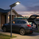 Outway T007 Nexa one-piece solar street light pole-mounted on a residential driveway at dusk, casting bright 1800lm white light over a parked SUV with surfboards on the roof rack. Woman loading boot under illuminated driveway. No cables visible — integrated LiFePO4 solar street light.