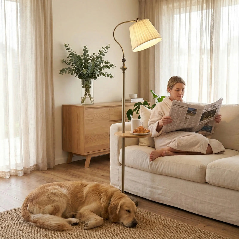 FL068 antique brass arc floor lamp with wood tray next to a cream sofa, warm white light, woman in bathrobe reading a newspaper with a golden retriever resting nearby