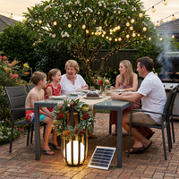 A family of five gathers around an outdoor dining table in a backyard patio setting, illuminated by warm string lights and a glowing solar-powered lantern. A small solar panel is visible on the brick ground next to the table.
