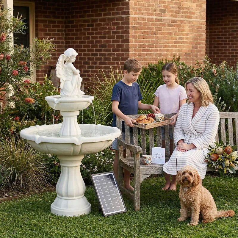 OUTWAY angel solar garden fountain on a lawn beside a wooden bench where two children present a breakfast tray to their mother in a striped robe with a golden retriever and Mother's Day card with solar panel visible on the grass