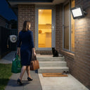E027EI Voro floodlight mounted above a brick front entrance at night, casting bright natural white light, woman arriving home with Woolworths bags, black cat on the doorstep