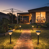 Four solar pathway lights illuminating a brick walkway in a landscaped backyard at dusk, with a house, patio, and yard garden visible in the background.