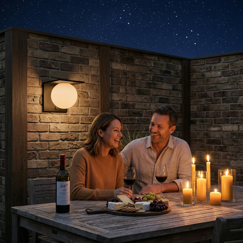 Couple laughing and sharing wine and cheese board at outdoor timber table on a starlit night, warm glow from Lumora Solar globe wall light mounted on brick wall behind them